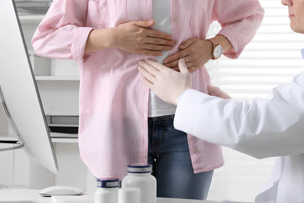 A healthcare professional wearing medical gloves performing a clinical abdominal palpation exam on a patient standing in a medical office.